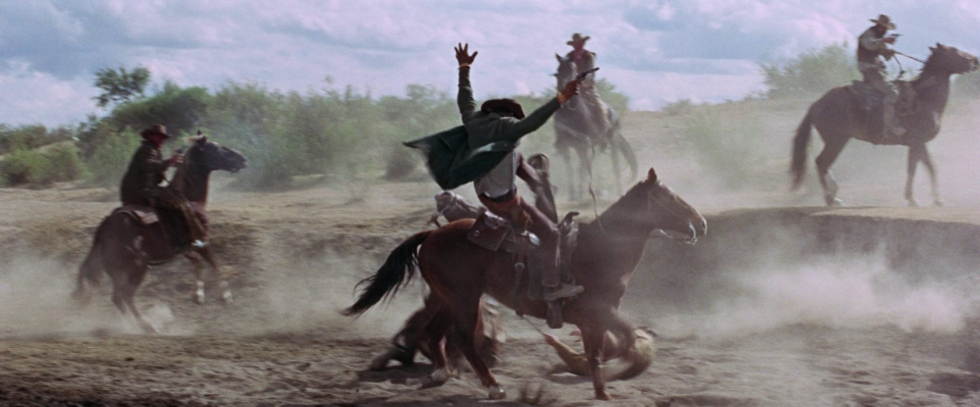 Schießerei zu Pferd, im Zentrum des Bildes ein Cowboy, der mit in die Luft gereckten Armen von seinem Pferd geschossen wird.
