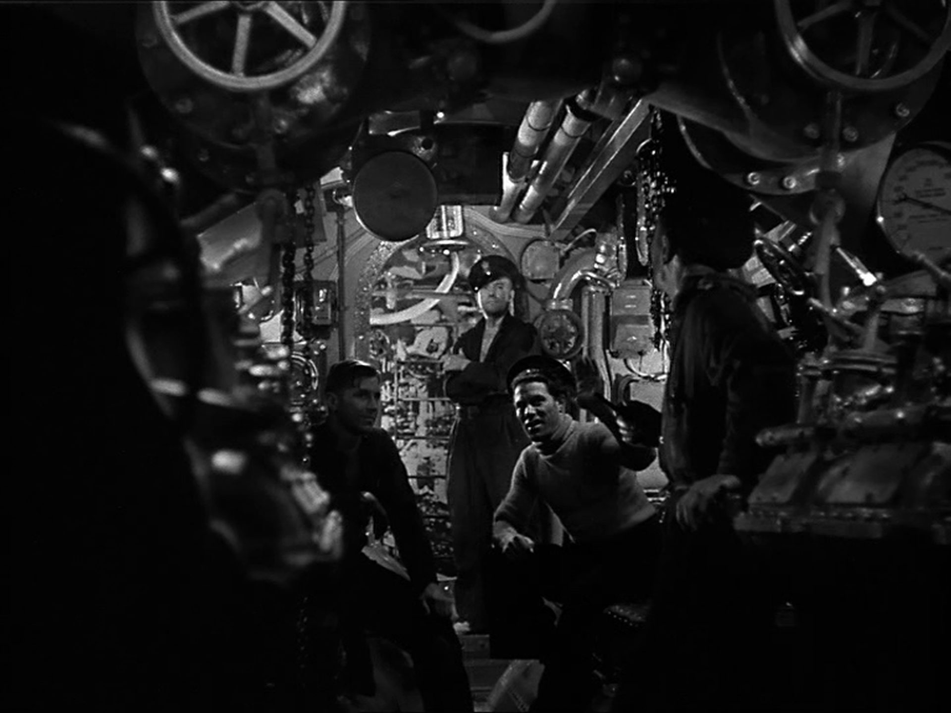 Black and white scene inside British submarine; view of three alert sailors surrounded by complex mechanics.