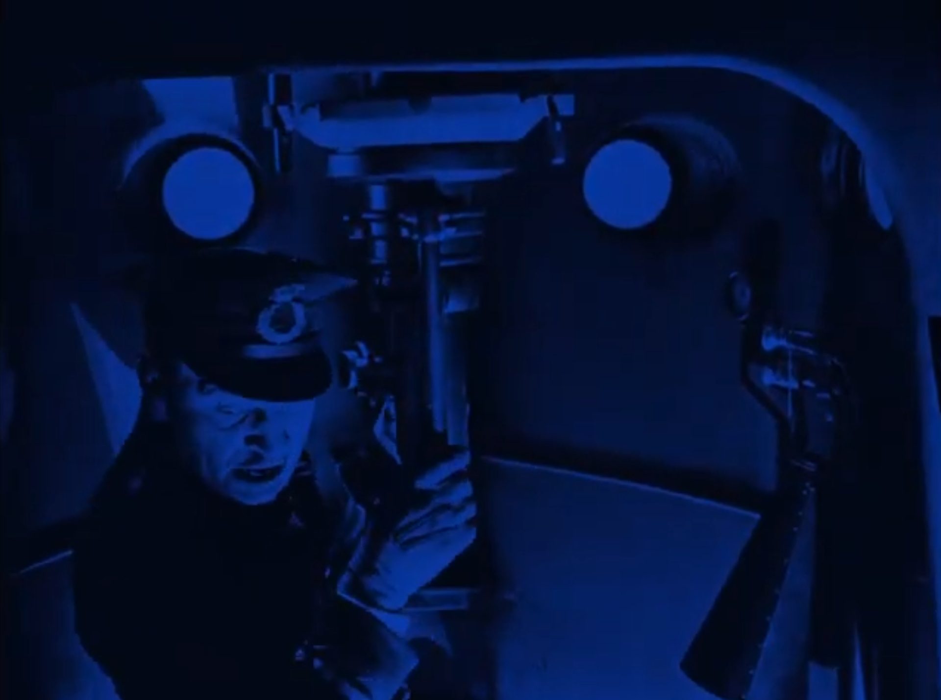 Blau eingefärbte Szene mit Wallace Beery als deutschem U-Boot-Kommandant am Periskop mit leicht aufgeregtem Blick nach schräg unten.