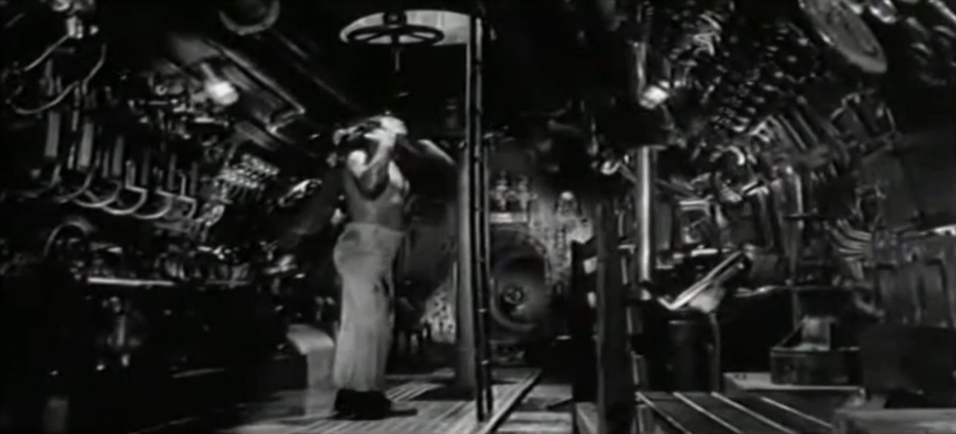 Schwarz-Weiß-Aufnahme vom Innern des italienischen U-Bootes mit seinen unzähligen Apparaturen; ein einzelner Seemann mit unbekleidetem Oberkörper steht unterhalb einer geöffneten Luke, durch ein wenig Tageslicht schimmert.