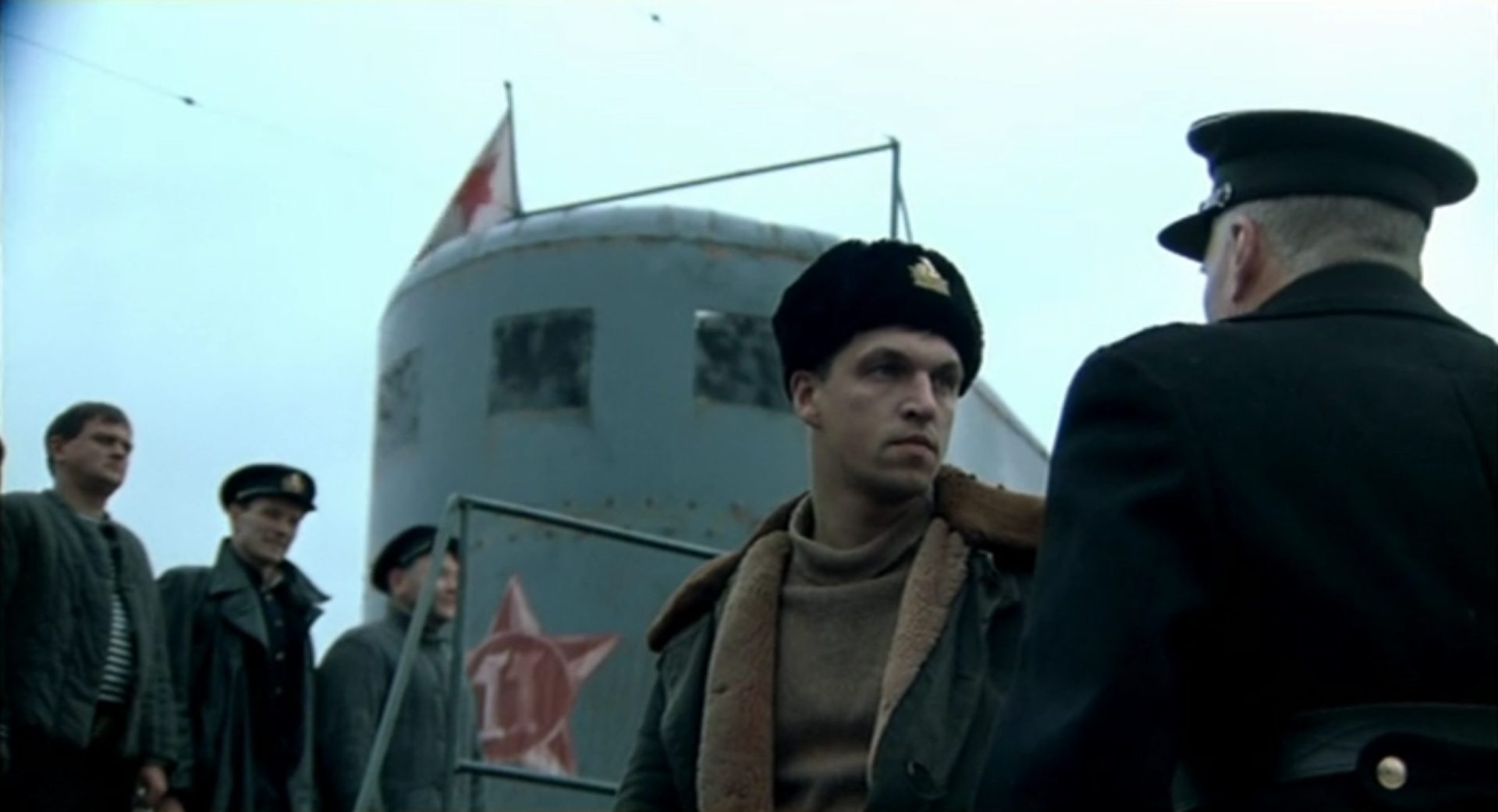 Dmitriy Orlov als sowjetischer U-Boot-Kommandant Aleksandr Marinin mit entschlossenem Blick, im Hintergrund der Turm seines U-Bootes und drei an Deck stehende Besatzungsmitglieder.