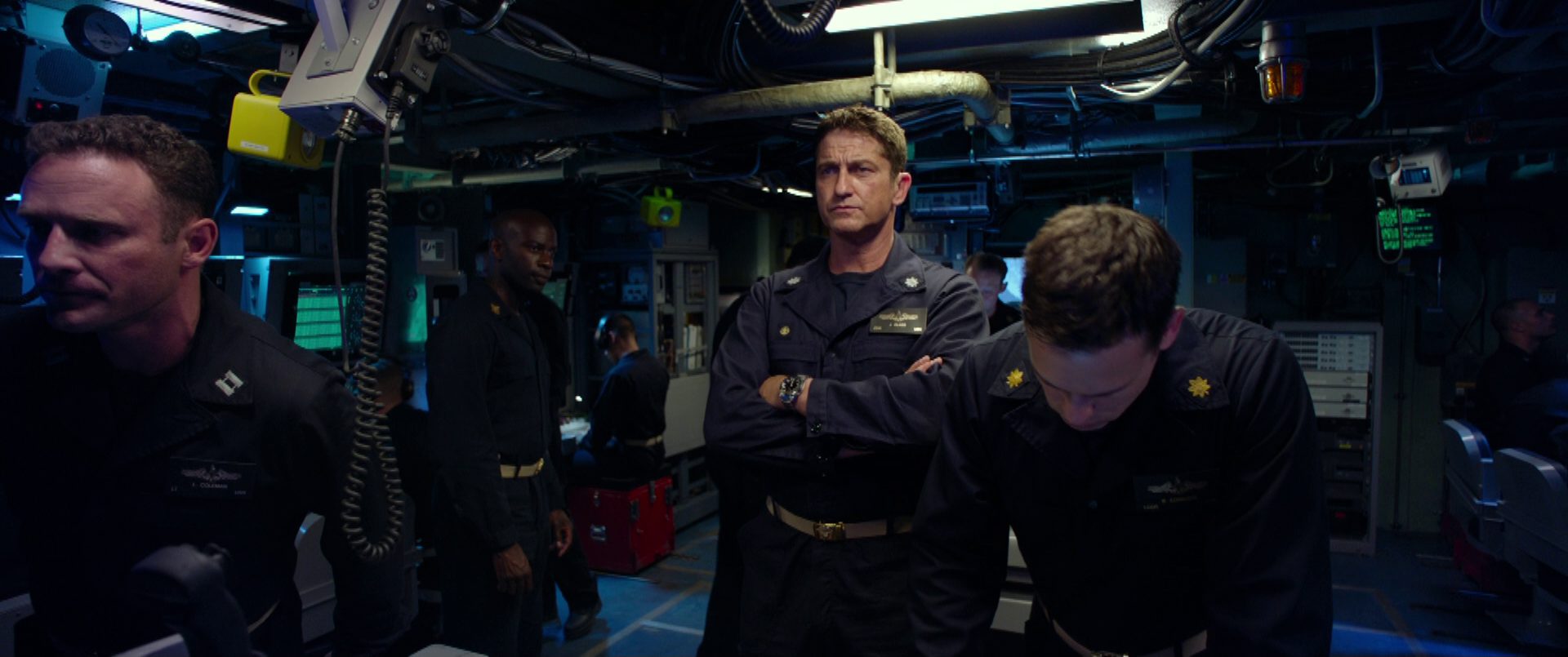 Gerard Butler as commander of a US Navy submarine with his arms crossed and his eyes focused and determined in the dimly lit control center in the presence of some of his sailors.