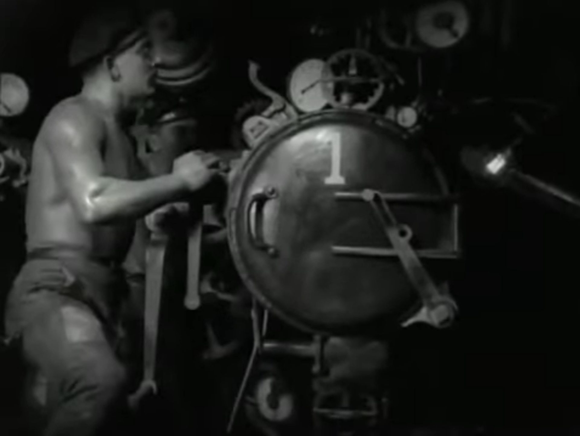 Schwarz-Weiß-Szene: Muskulöser Seemann mit nacktem Oberkörper am Torpedorohr, auf dessen Luke in weißer Serifenschrift eine 1 aufgebracht ist.