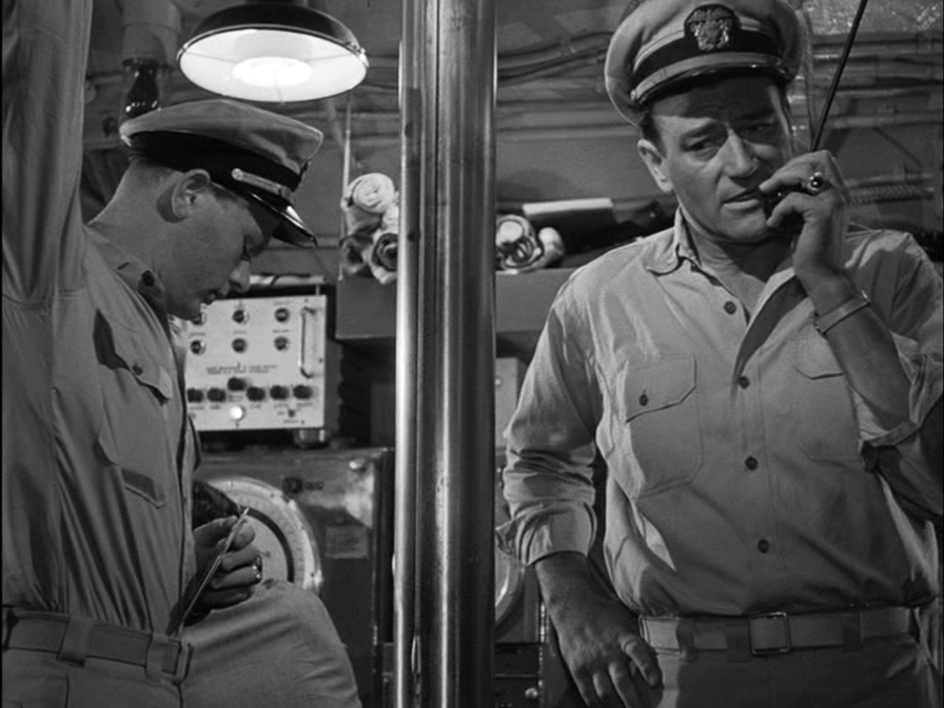 Schwarz-weiße Nahaufnahme von John Wayne als U-Boot-Kommandant am Bordmikrofon, neben ihm, getrennt durch das Periskop, ein Seemann.