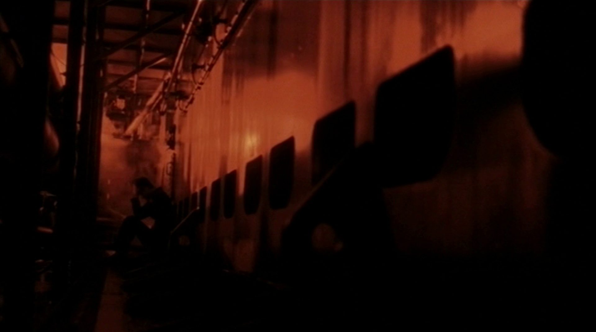 Gloomy, reddish colored scene in the submarine, in the background a sailor crouches, his face buried in his hands.