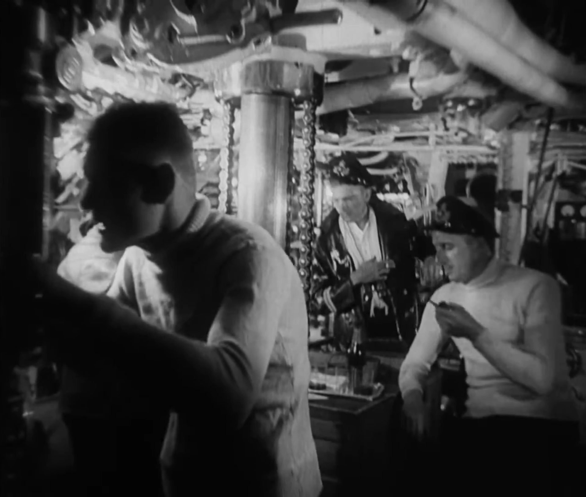 Schwarz-Weiß-Szene in der Kommandozentrale des deutschen U-Bootes; zwei Crewmitglieder und der Kapitän im weißen Rollkragenpullover mit Pfeife.