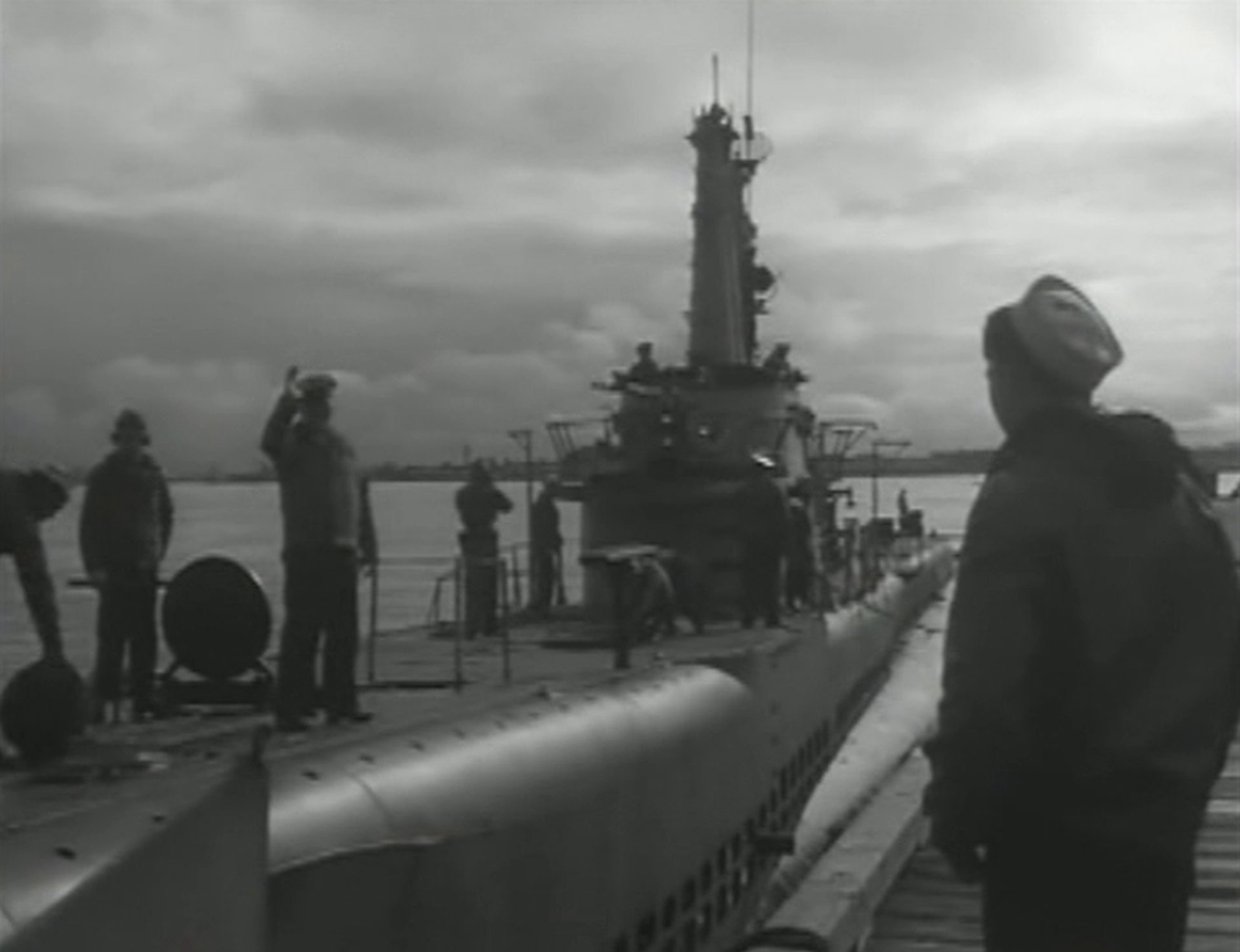 Schwarz-Weiß-Aufnahme eines Navy-U-Bootes beim Auslaufen, an Deck mehrere Seeleute, von denen einer winkt, am rechten Bildrand ein Matrose am Pier.