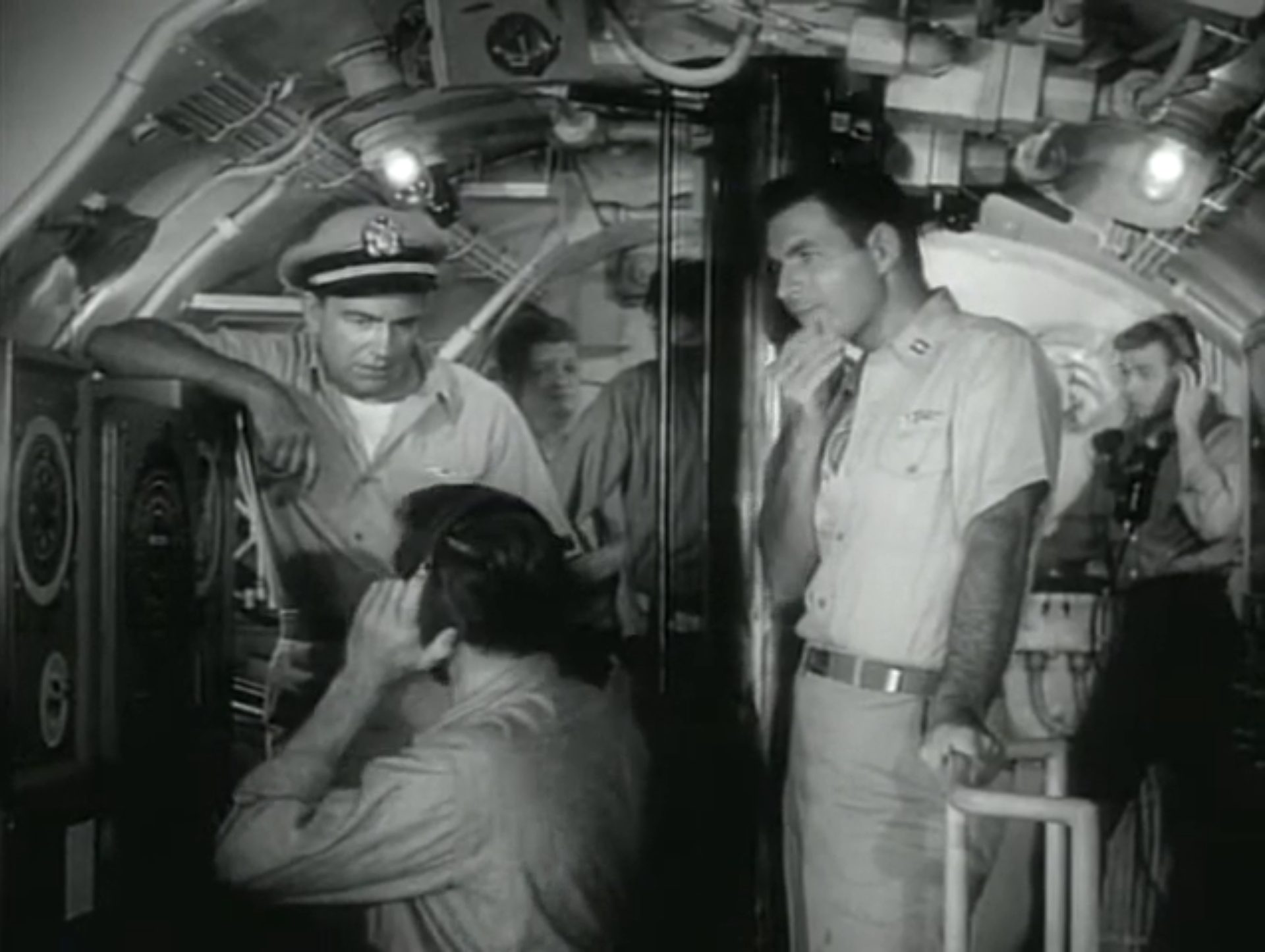 Schwarz-Weiß-Aufnahme in der Kommandozentrale eines Navy-U-Bootes; der Kapitän lehnt an einem Gerät und blickt zum davor sitzenden Seemann herab, der Kopfhörer trägt; in der Mitte steht ein Offizier in nachdenklicher Pose neben dem Periskop, im Hintergrund weitere Seeleute.