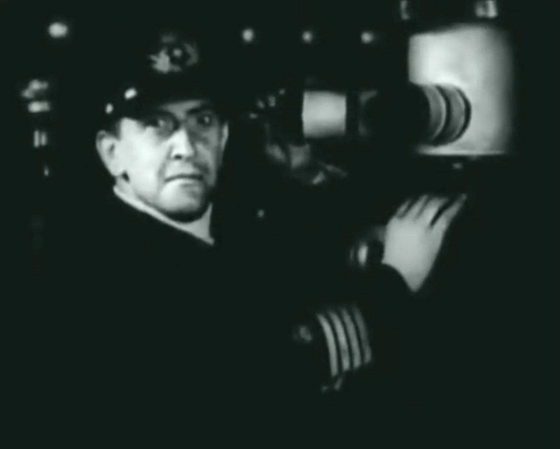 Schwarz-weiße Nahaufnahme eines deutschen U-Boot-Kapitäns am Periskop, der in die Kamera blickt.