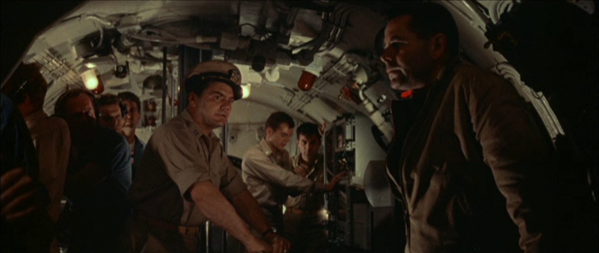 Blick in die Kommandozentrale des Navy-U-Bootes, in der Mitte Ernest Borgnine als Erster Offizier mit besorgtem Blick zum Kapitän, der düster ausgeleuchtet in nachdenklicher Haltung am Rand lehnt und von Glenn Ford gespielt wird; im Hintergrund mehrere Seeleute.