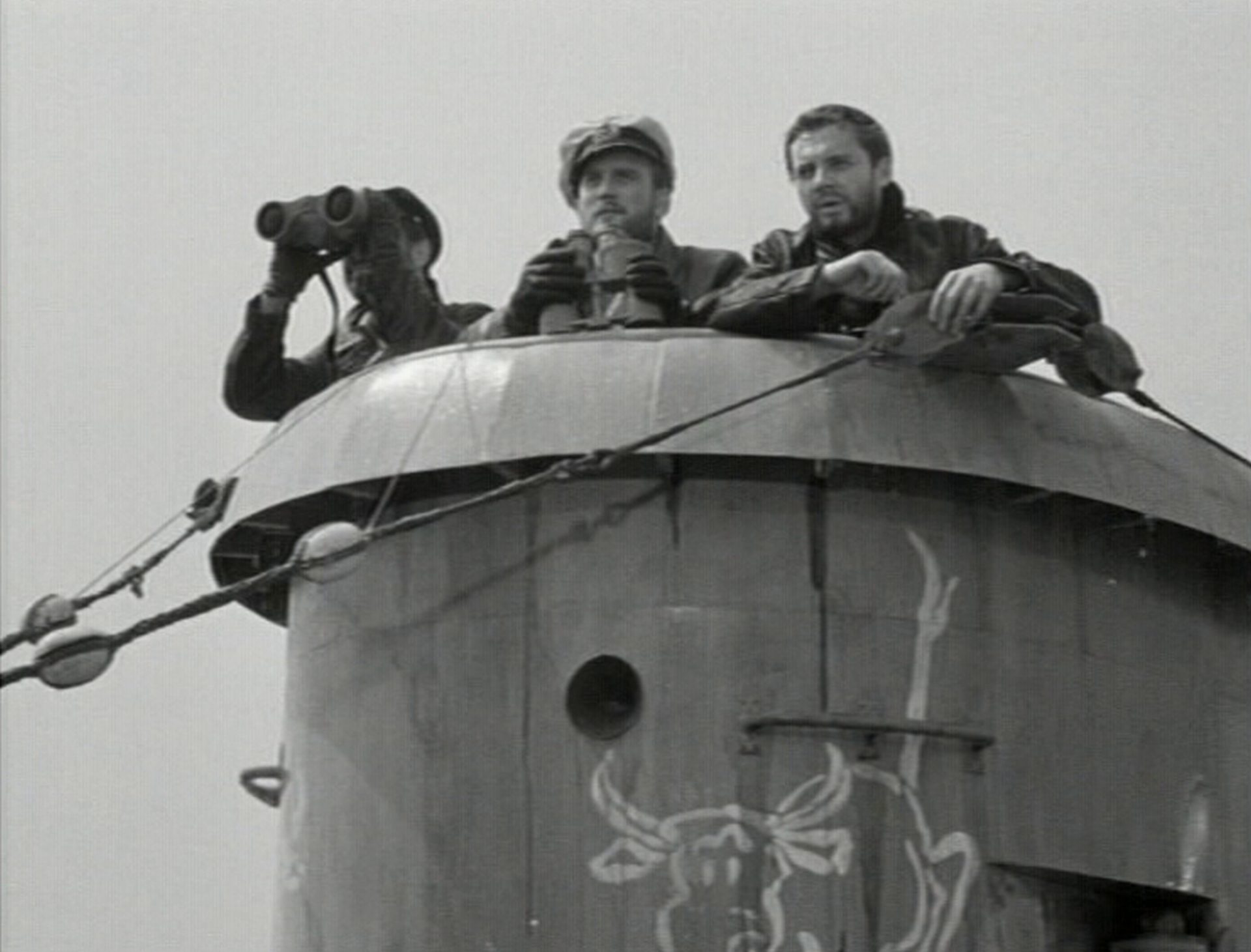 Schwarz-Weiß-Aufnahme von Dieter Eppler als U-Boot-Kommandant Günther Prien im Ausguck der U-47 mit abgesetztem Fernglas, zu beiden Seiten jeweils einer seiner Seeleute, einer davon mit angesetztem Fernglas; der Turm trägt einen mit weißen Konturen aufgemalten Stier als Maskottchen.