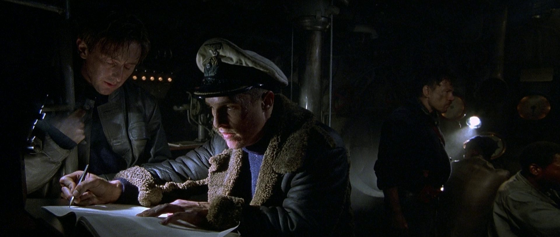 In a dimly lit environment, Matthew McConaughey, as an American commander in German uniform, concentrates in front of an open notebook in which he is writing in the presence of a comrade, with three other submariners in the background.