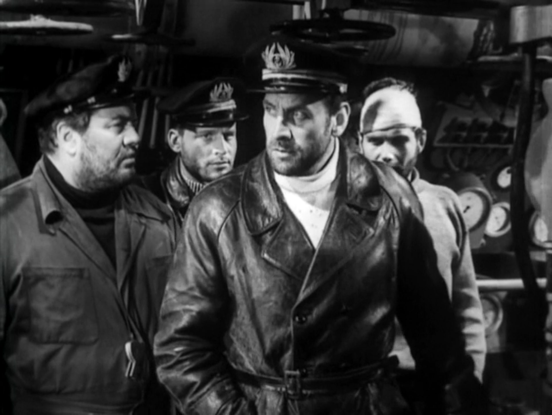 Black and white scene of the sailors cooped up in the sunken submarine; the captain, dressed in a leather coat, looks skeptically to the side, one of the sailors is wearing a white bandage.