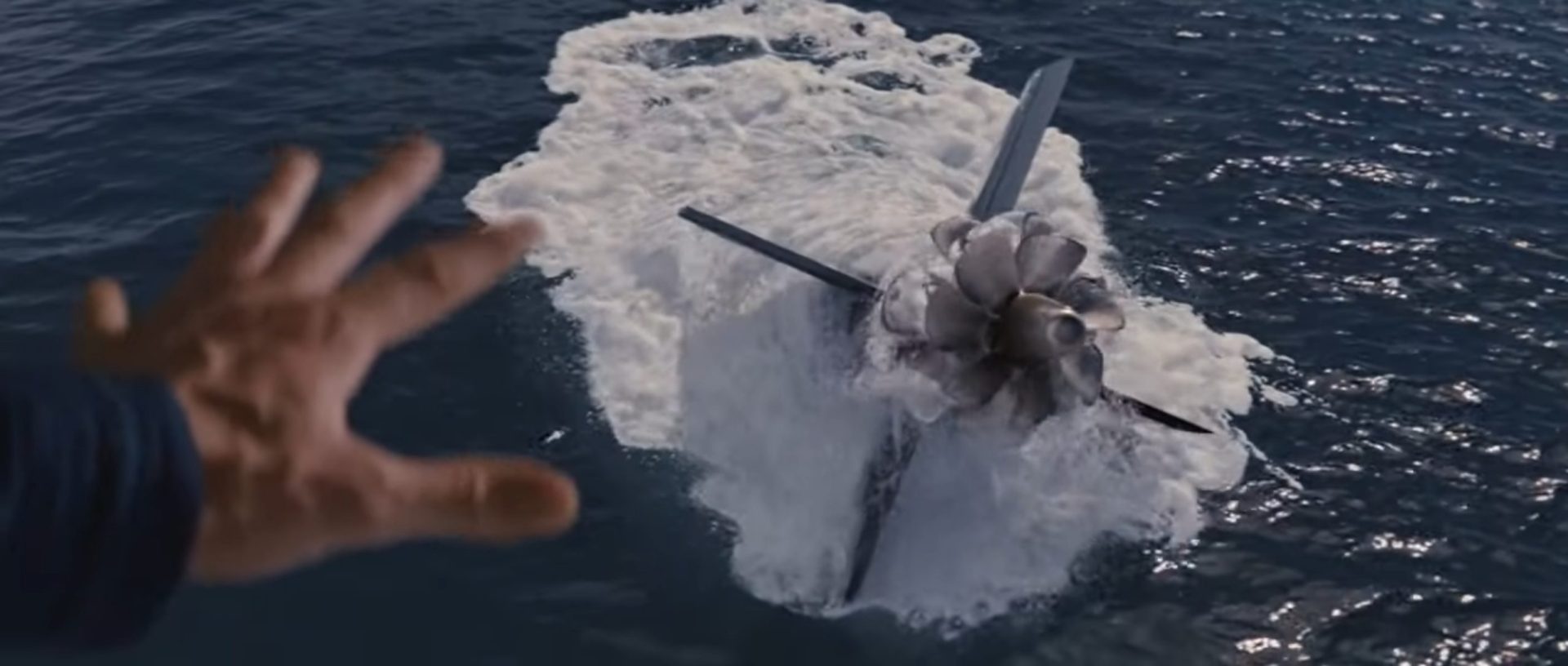 Im Vordergrund eine ausgestreckte Hand, weiter unten im ragt das Heck eines U-Bootes aus der Meeresoberfläche heraus.