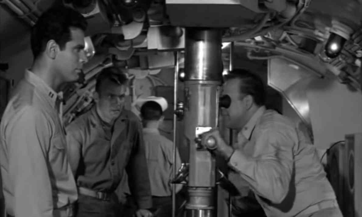 Schwarz-Weiß-Szene mit dem Kommandanten beim Blick durch das Sehrohr, zwei Offiziere haben ihre Augen auf ihn gerichtet.