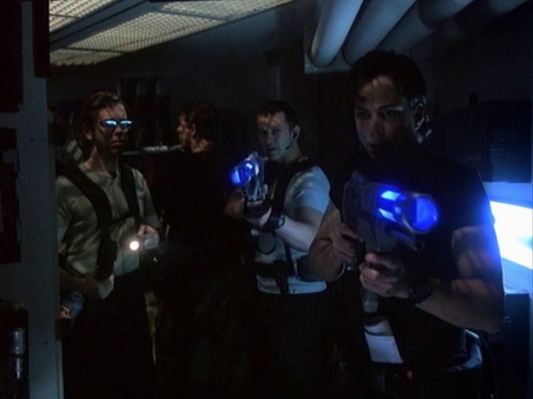 Vier Männer in einem düsteren Korridor des U-Bootes, zwei von ihnen mit futuristischen Waffen im Anschlag, die blau leuchten.