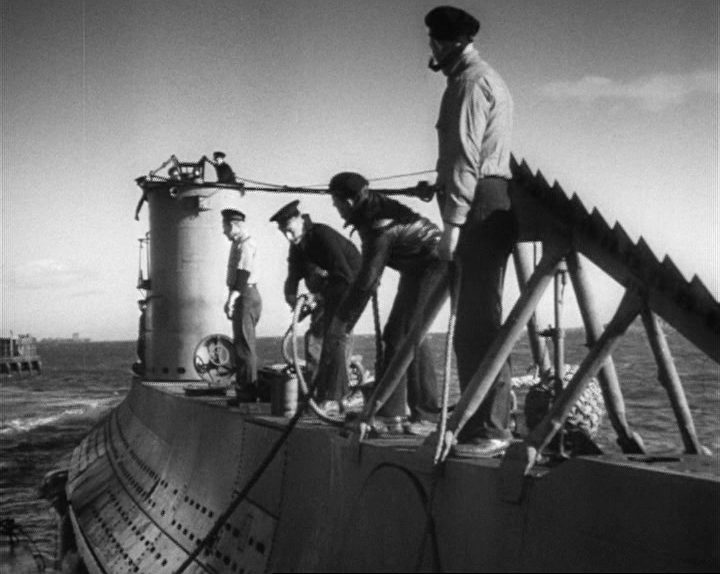 Schwarz-weiße Nahaufnahme von Männern an Deck eines ablegenden U-Bootes, im Vordergrund die Netzsäge am Bug.