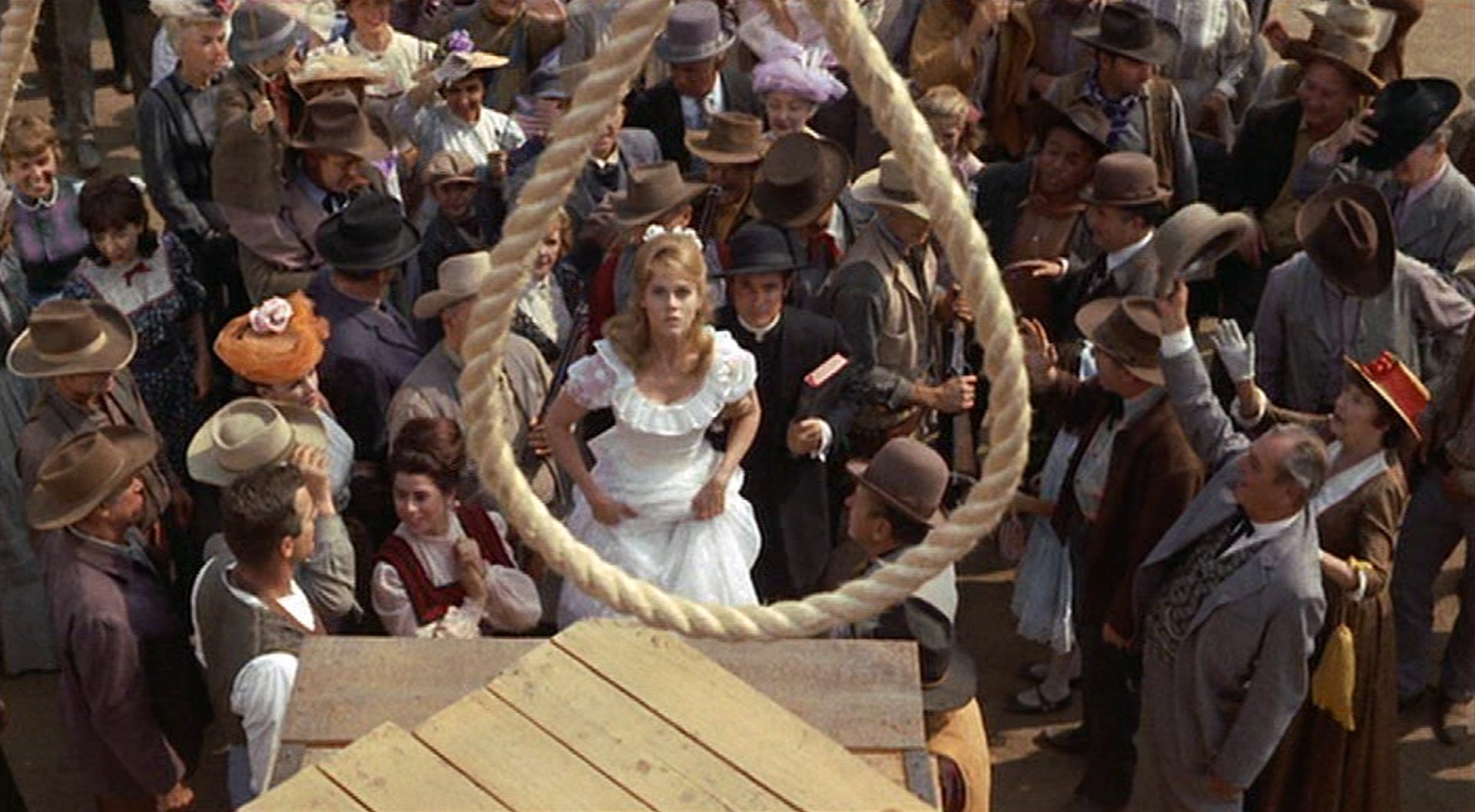 View through gallows noose of Cat Ballou in white dress amidst lynching audience.