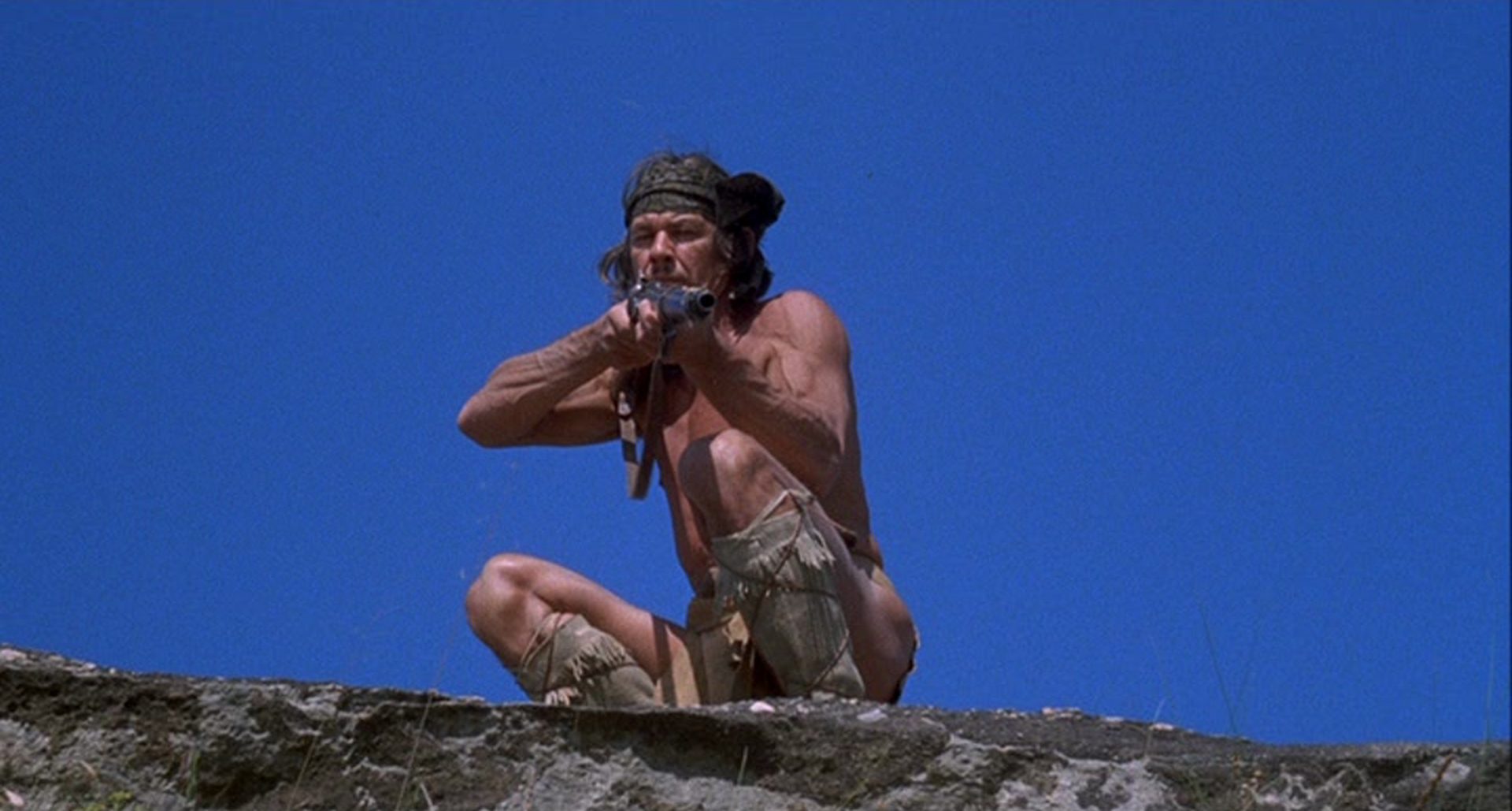 Charles Bronson as Chato against a blue sky in an alert fighting pose and with rifle at the ready.
