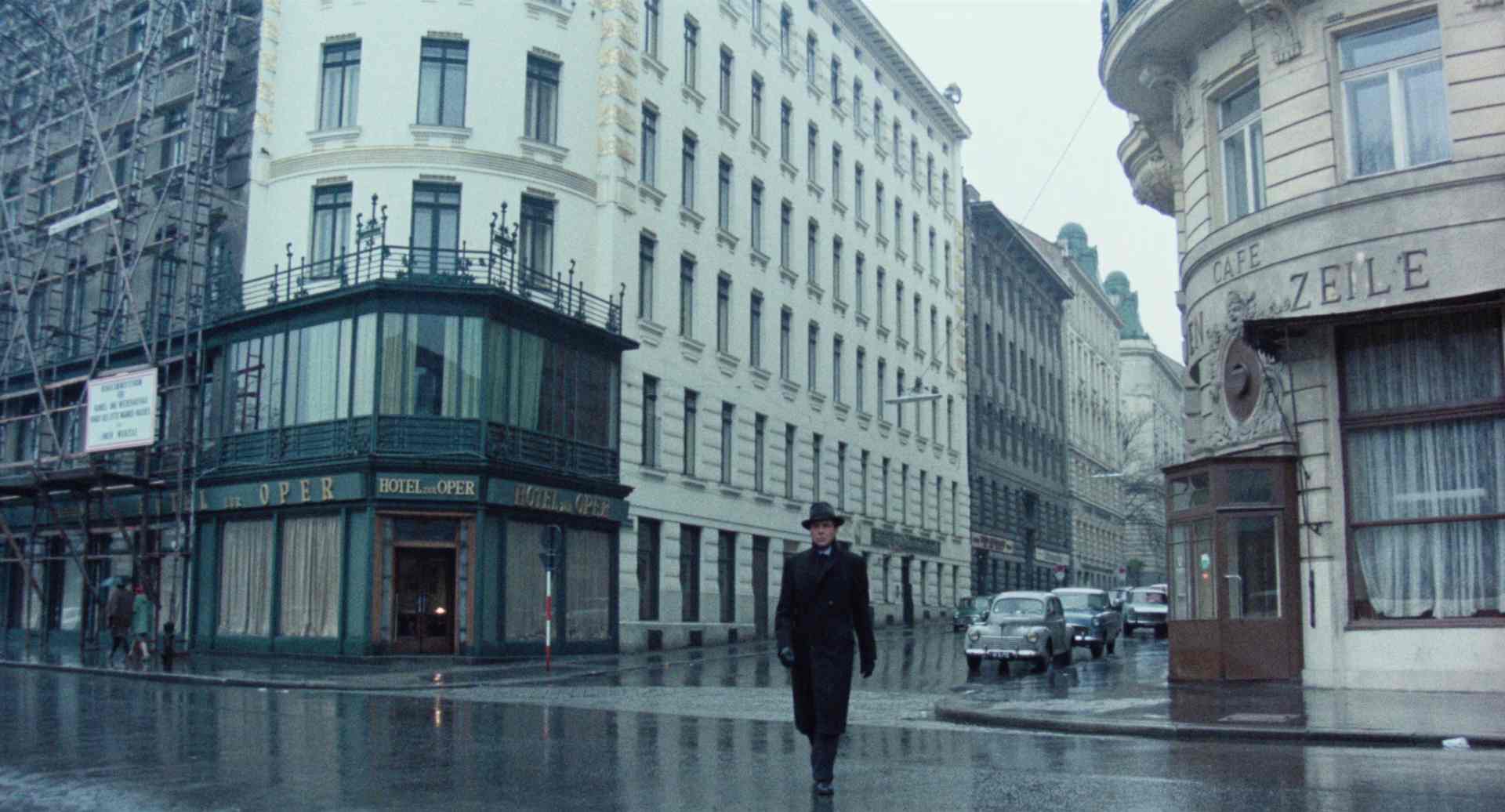 Dirk Bogarde als Portier im Mantel mit Hut auf der verregneten Straße vor dem Hotelgebäude, das an der linken Seite im Bild eingerüstet ist.