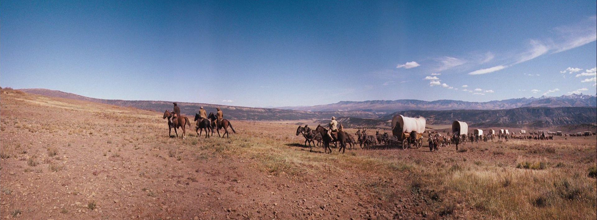 Ein Siedlertreck mit seinen Planwagen unterwegs durch die Prärie vor strahlend blauem Himmel.
