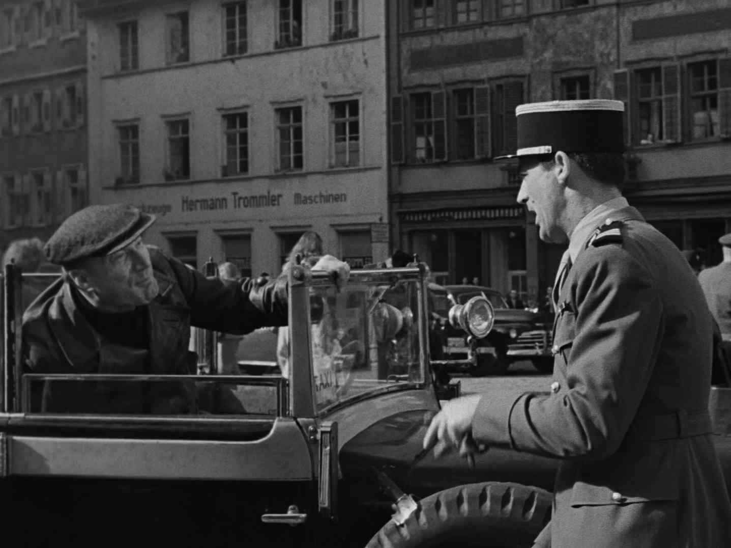 Cary Grant als französischer Offizier im Gespräch mit seinem Fahrer, der im offenen Wagen sitzt; im Hintergrund steht an der Fassade eines Hauses „Hermann Trommler Maschinen“.