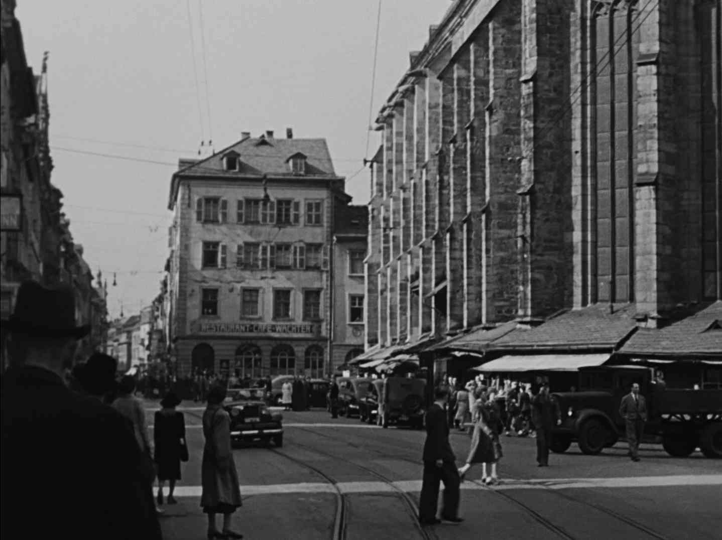 Schwarz-Weiß-Aufnahme vom Marktplatz in Heidelberg; rechts ist die Außenwand der Heiliggeistkirche zu erkennen, im Hintergrund ein Eckhaus mit einem Schild, das die Aufschrift „Restaurant Cafe Wachter“ in Großbuchstaben trägt.