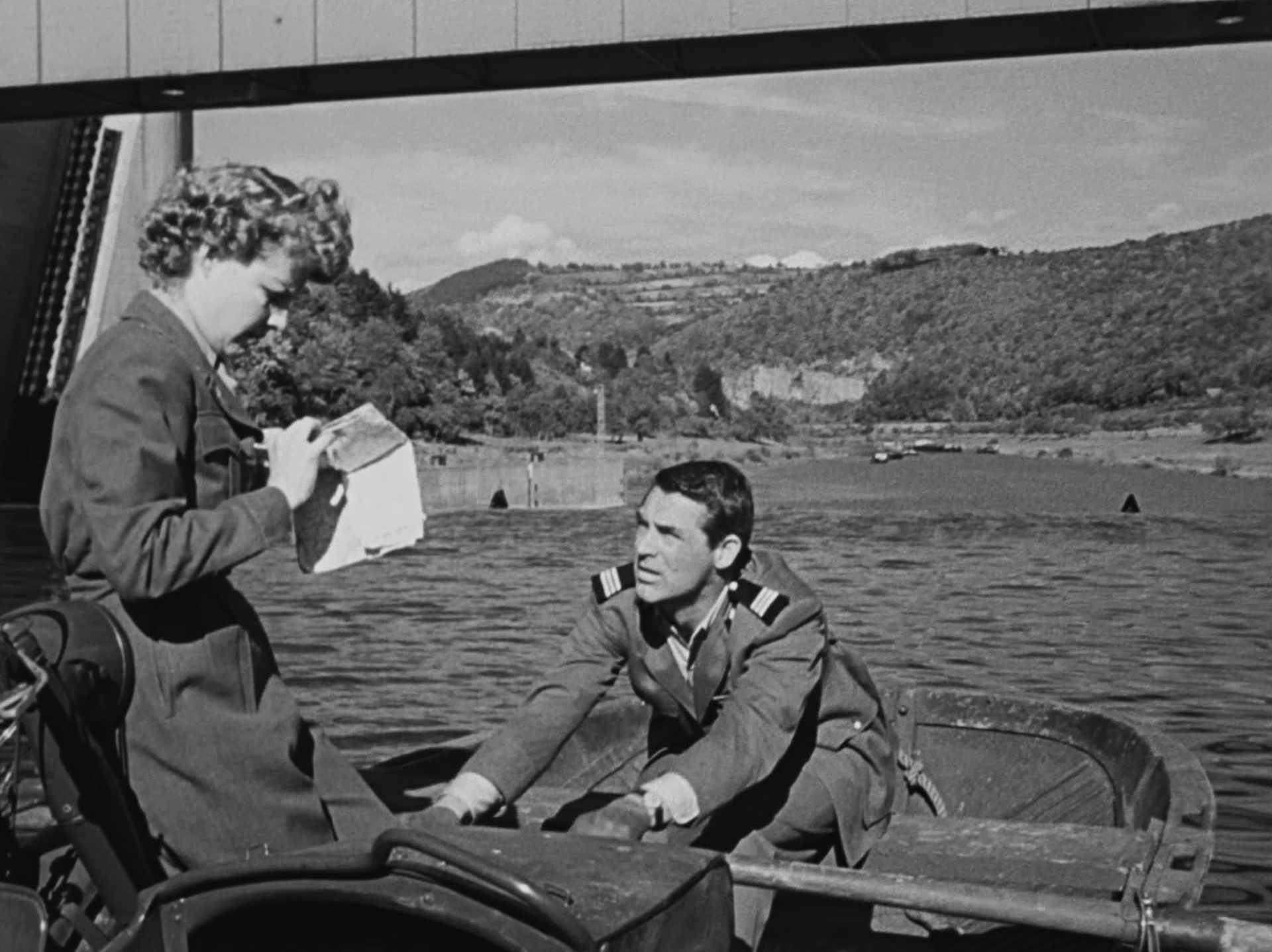 Schwarz-Weiß-Szene: Ann Sheridan und Cary Grant in einem Ruderboot vor der Staustufe Rockenau, kurz vor dem Abgrund, im Hintergrund erstrecken sich die Hänge großer Hügelketten; sie schaut auf eine Faltkarte, er rudert.