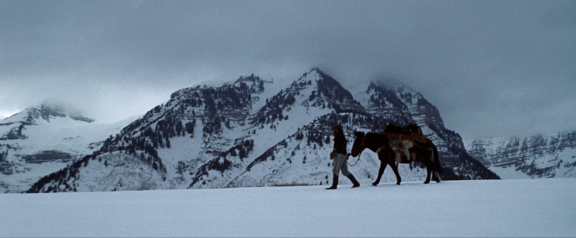 Robert Redford, as Jeremiah Johnson, trudges through an icy landscape with a horse on a leash and snow-covered mountains in the background.