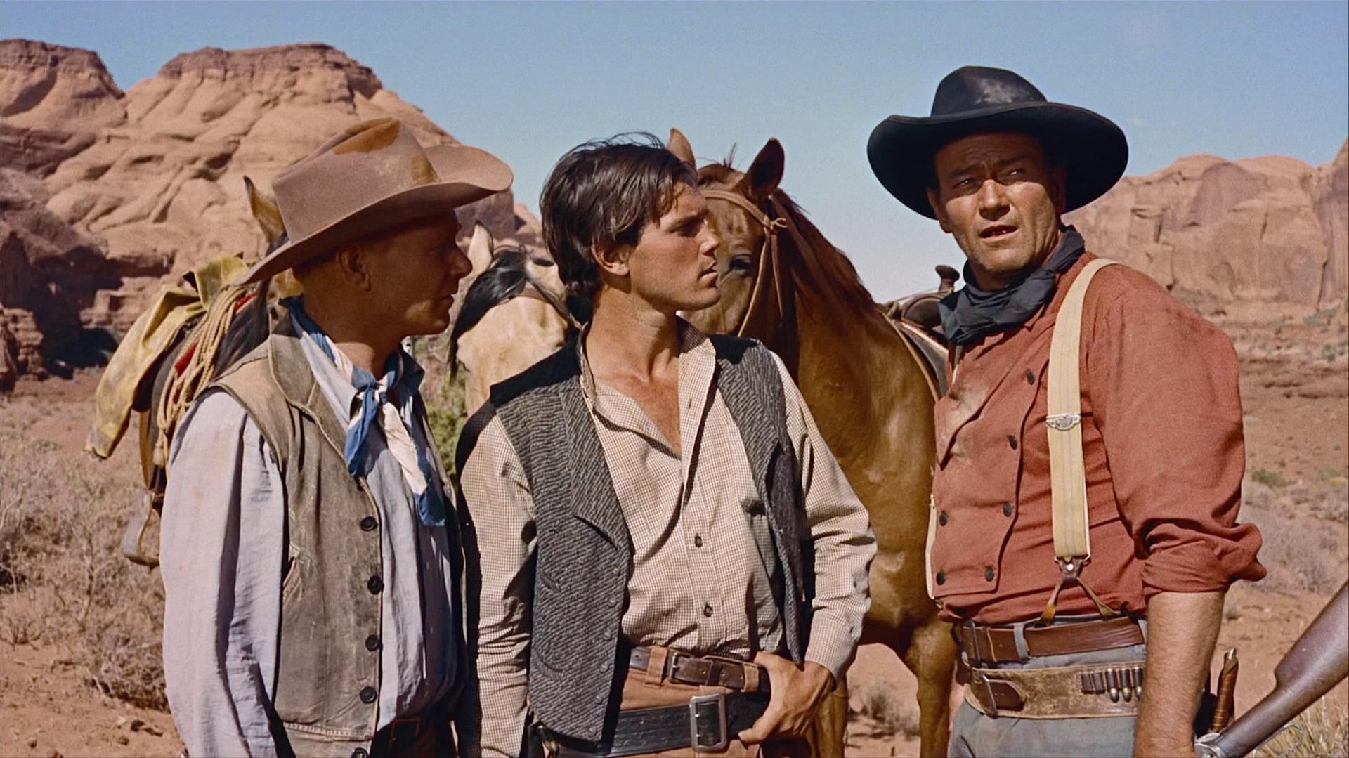 John Wayne als Ethan Edwards mit zwei Kompagnons bei warmem Klima in der Wüste.