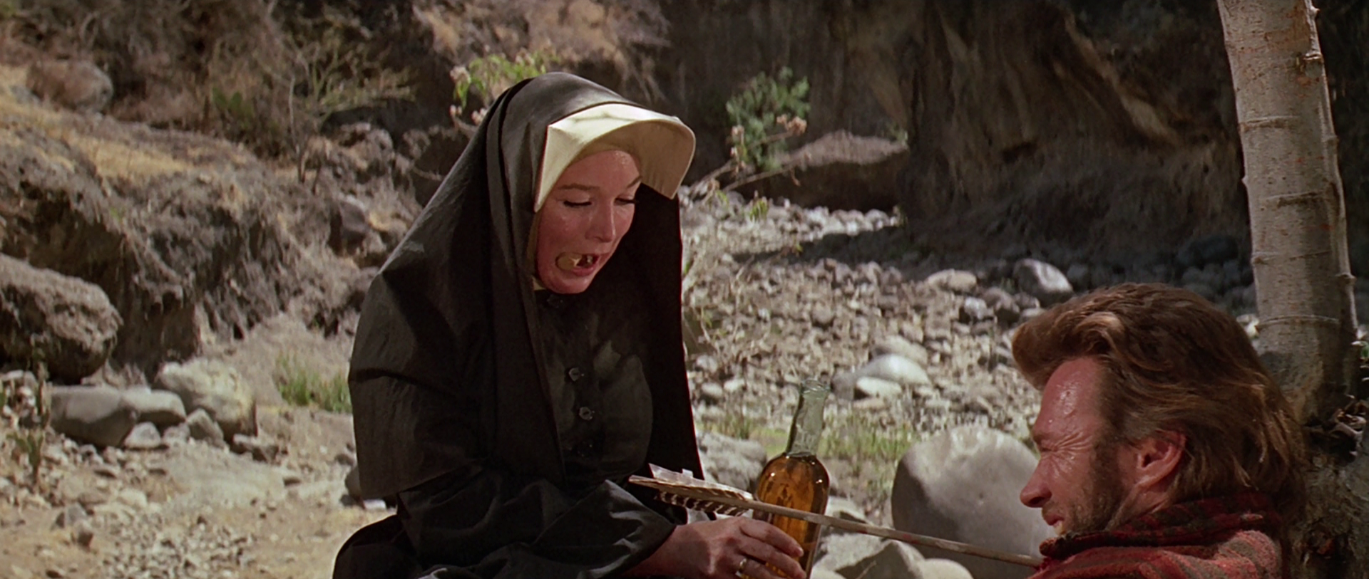 Sister Sara has a cork between her lips and hands a bottle of whiskey to Hogan, who has been wounded by an arrow.