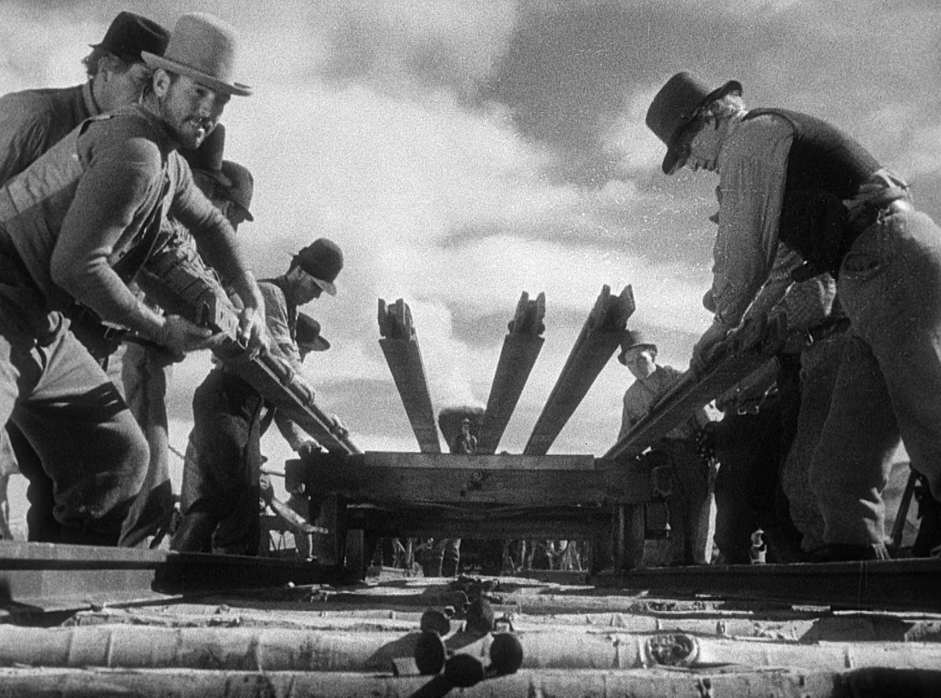 Schwarz-Weiß-Szene aus der Froschperspektive aus dem Gleisbett: Mehrere Arbeiter hieven Schienen von einem flachen Waggon.