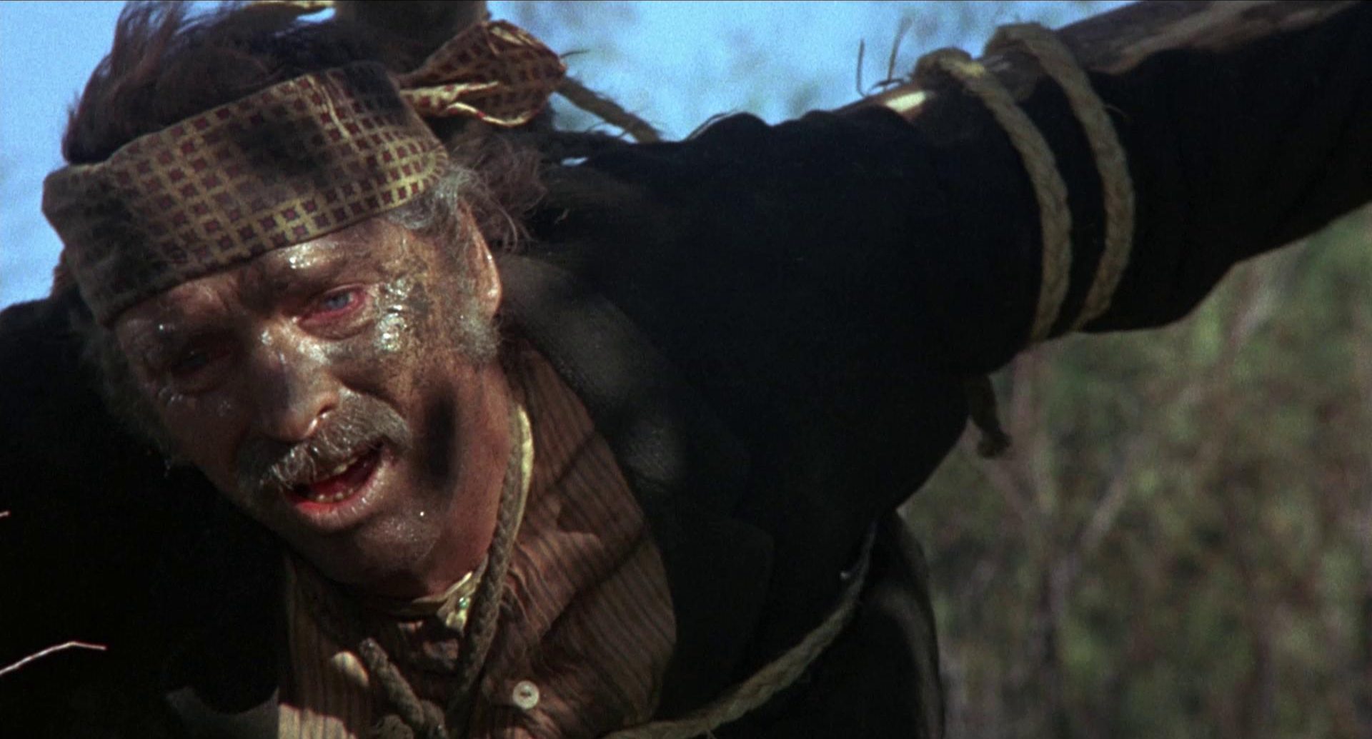 Nahaufnahme von Burt Lancaster als Valdez, der mit strapaziös-ramponiertem Gesicht an ein Holzkreuz gefesselt durch die Wildnis torkelt.
