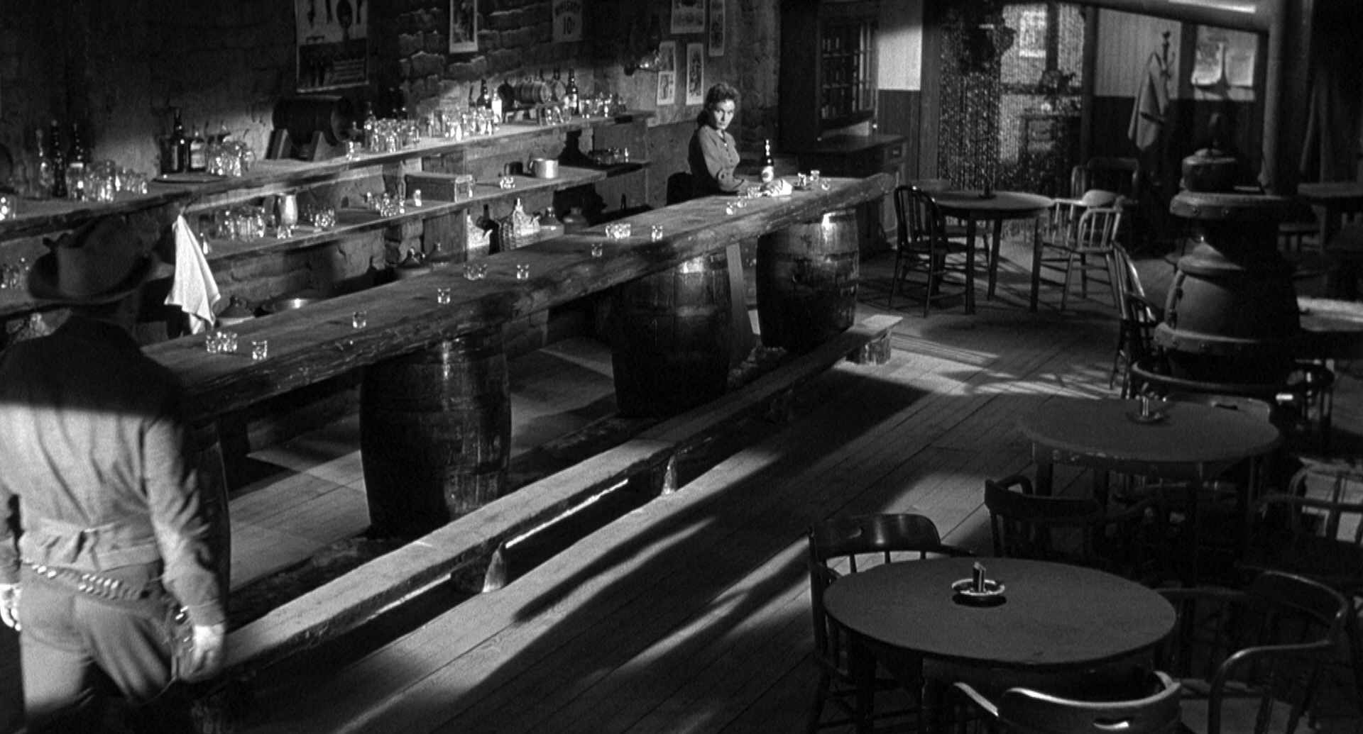 Expressionistic black and white scene in which a cowboy enters a saloon empty except for the bartender; you can’t see his face, she is looking at him from across the room.