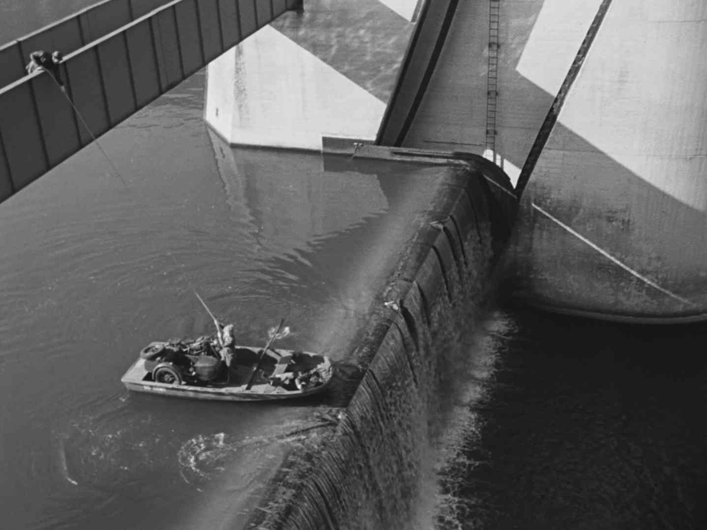 Schwarz-Weiß-Szene, in der sich ein kleines, flaches Boot kurz vor dem Abgrund an einem Wehr befindet; von einer Brücke hat jemand ein Rettungsseil geworfen, das ein Bootspassagier gerade strafft.