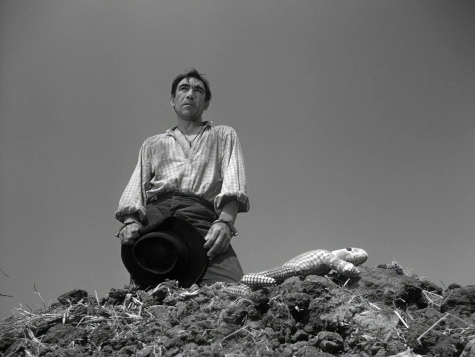 Black-and-white scene in which a jaded Anthony Quinn can be seen looking pensively into the distance from a frog’s-eye view of a burial mound on which a doll is lying.