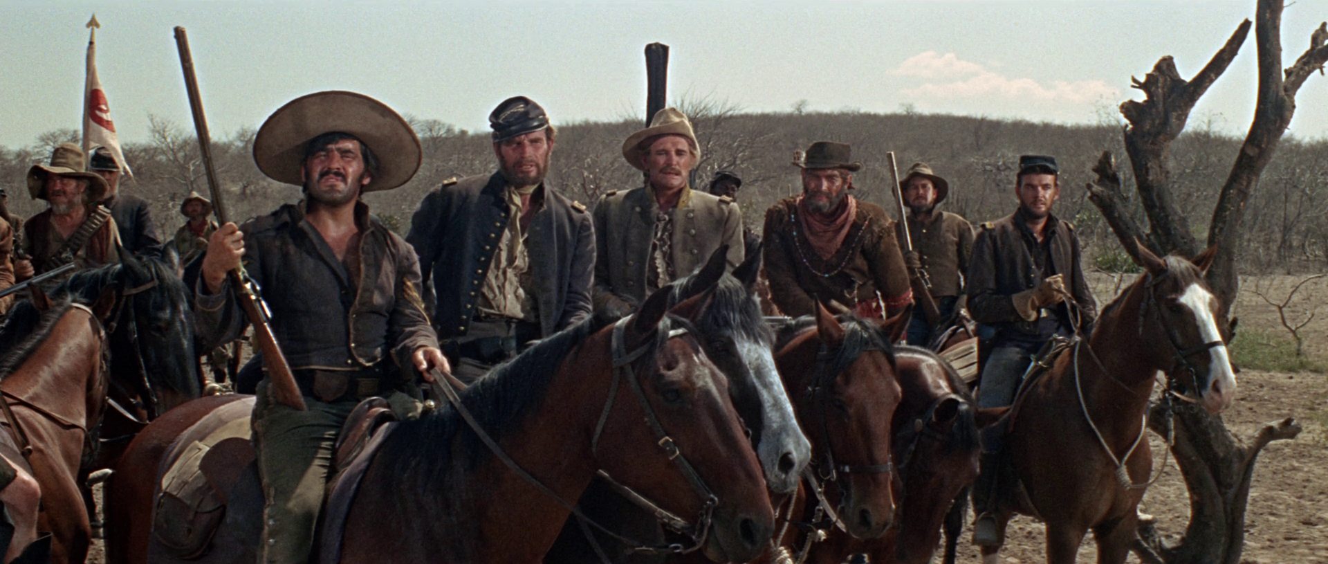 Mario Adorf, Charlton Heston, Richard Harris, James Coburn und andere als Kavalleristen zu Pferd in trist-trockener Umgebung.