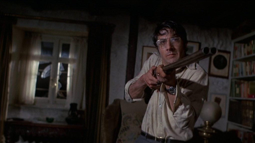 Frontal shot of Dustin Hoffman as David Sumner in the gloomy living room of his English country house in a concentrated defensive posture with a double-barreled rifle at the ready.