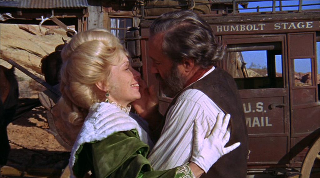 Stella Stevens as Hildy in noble garb and Jason Robards as Cable Hogue in businesslike outfit; both stand intimately against the backdrop of a stagecoach.