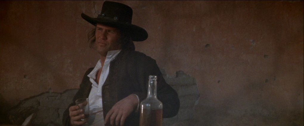 Kris Kristofferson as Billy the Kid in a casual pose in front of a wall with a whiskey glass in his hand, a half-full whiskey bottle is in the foreground.