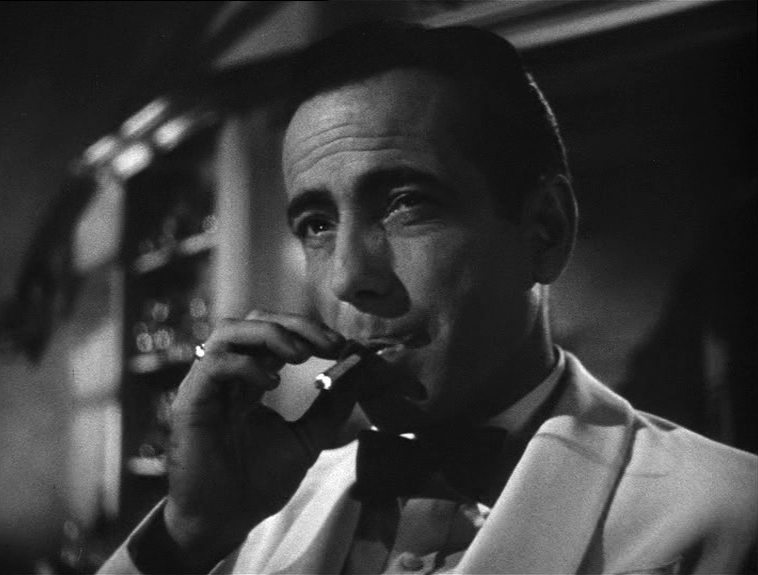 Black and white close-up of Humphrey Bogart as Rick Blaine, taking a drag on his glowing cigarette in a white suit.