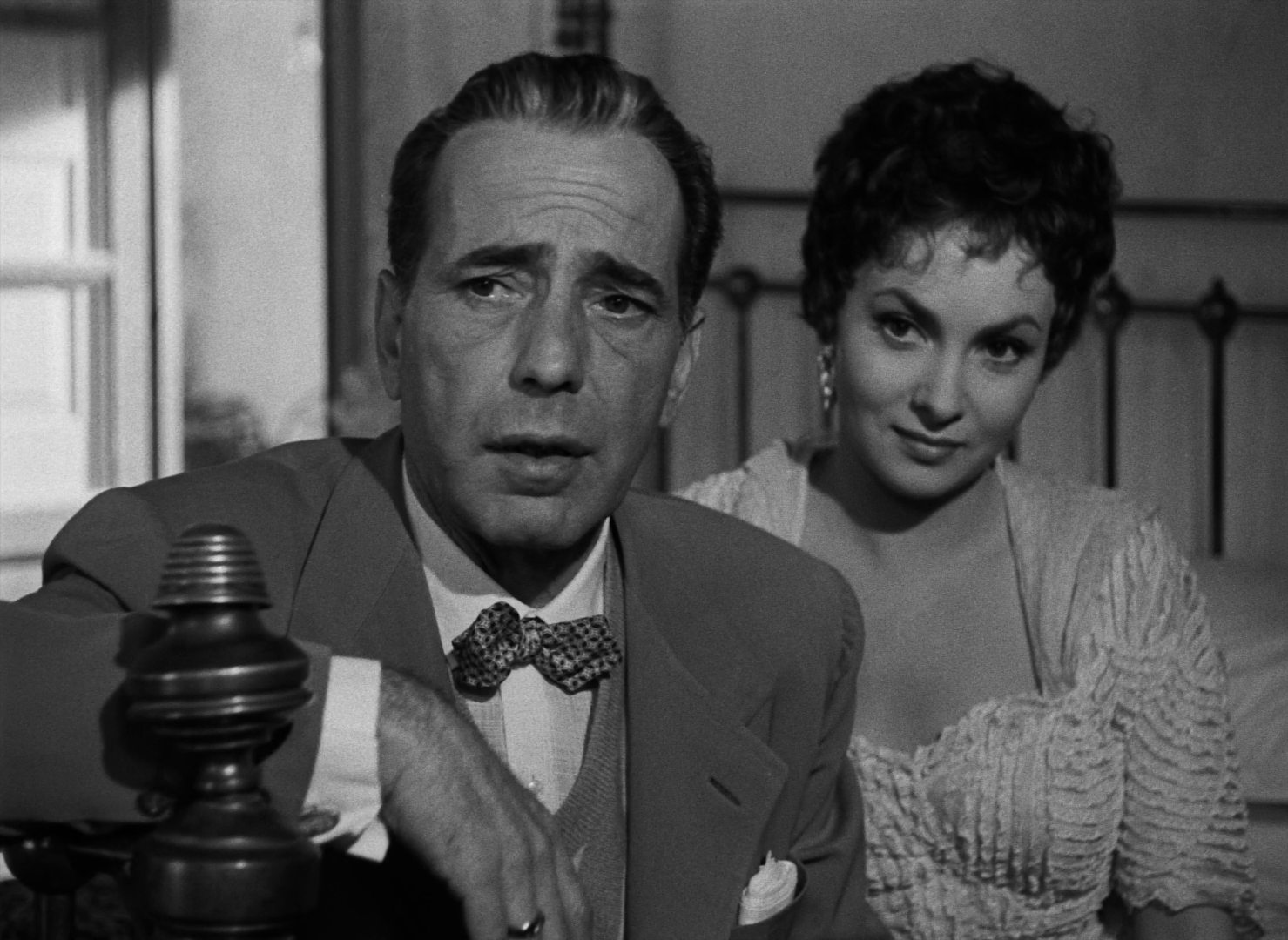 Black and white close-up of Humphrey Bogart and Gina Lollobrigida: both sitting on a hotel bed, elegantly dressed, with attentive looks.