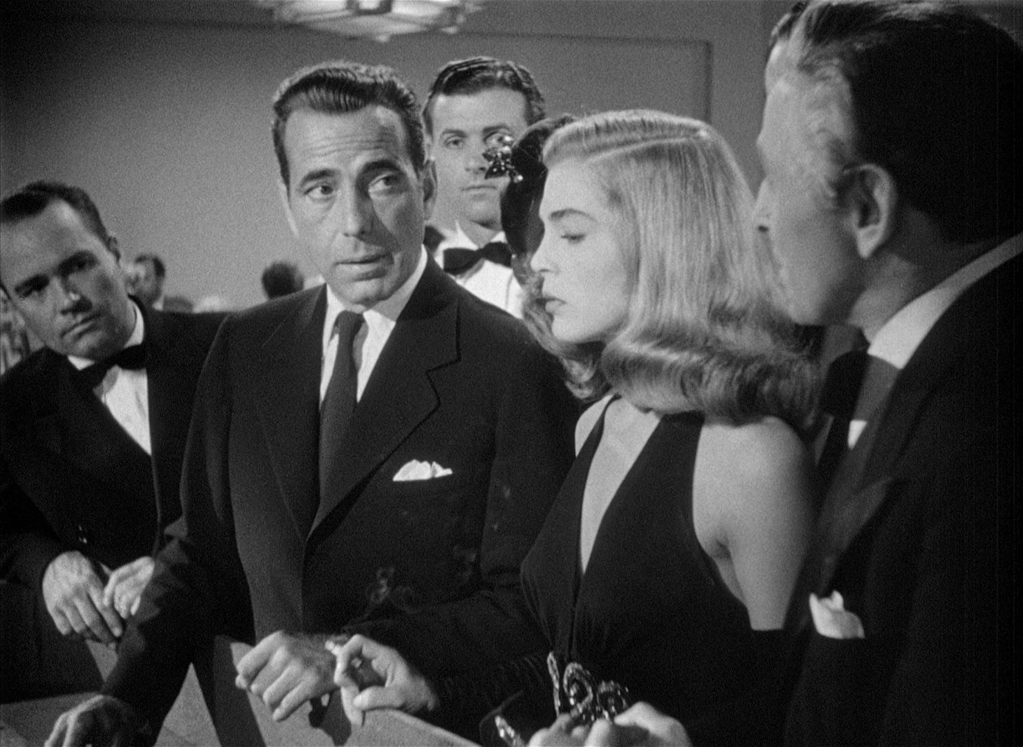 Black-and-white scene: Humphrey Bogart at the gambling table, attentively observing a man in the foreground, next to Lizabeth Scott, all dressed in evening wear.