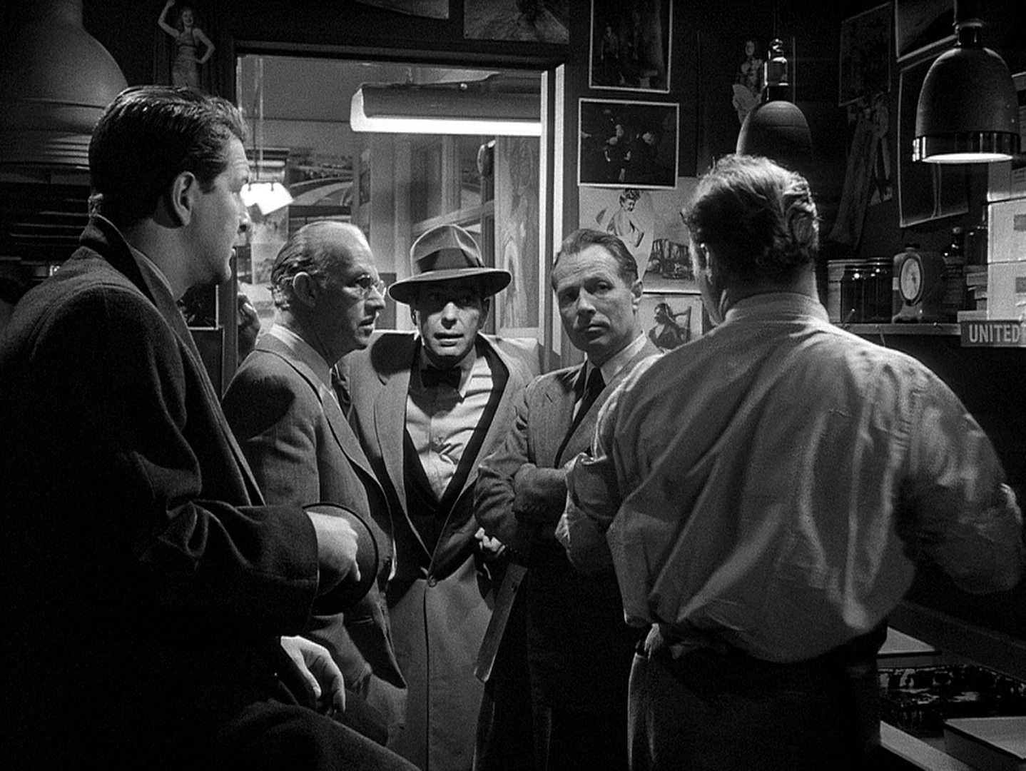 Schwarz-Weiß-Szene: Humphrey Bogart in Noir-Pose mit Mantel und Hut als Chefredakteur in einem kleinen Redaktionsbüro im Gespräch mit vier Mitarbeitern.