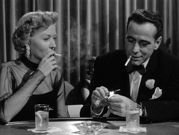 Black and white scene with Gloria Grahame and Humphrey Bogart having a cigarette romance in a bar.