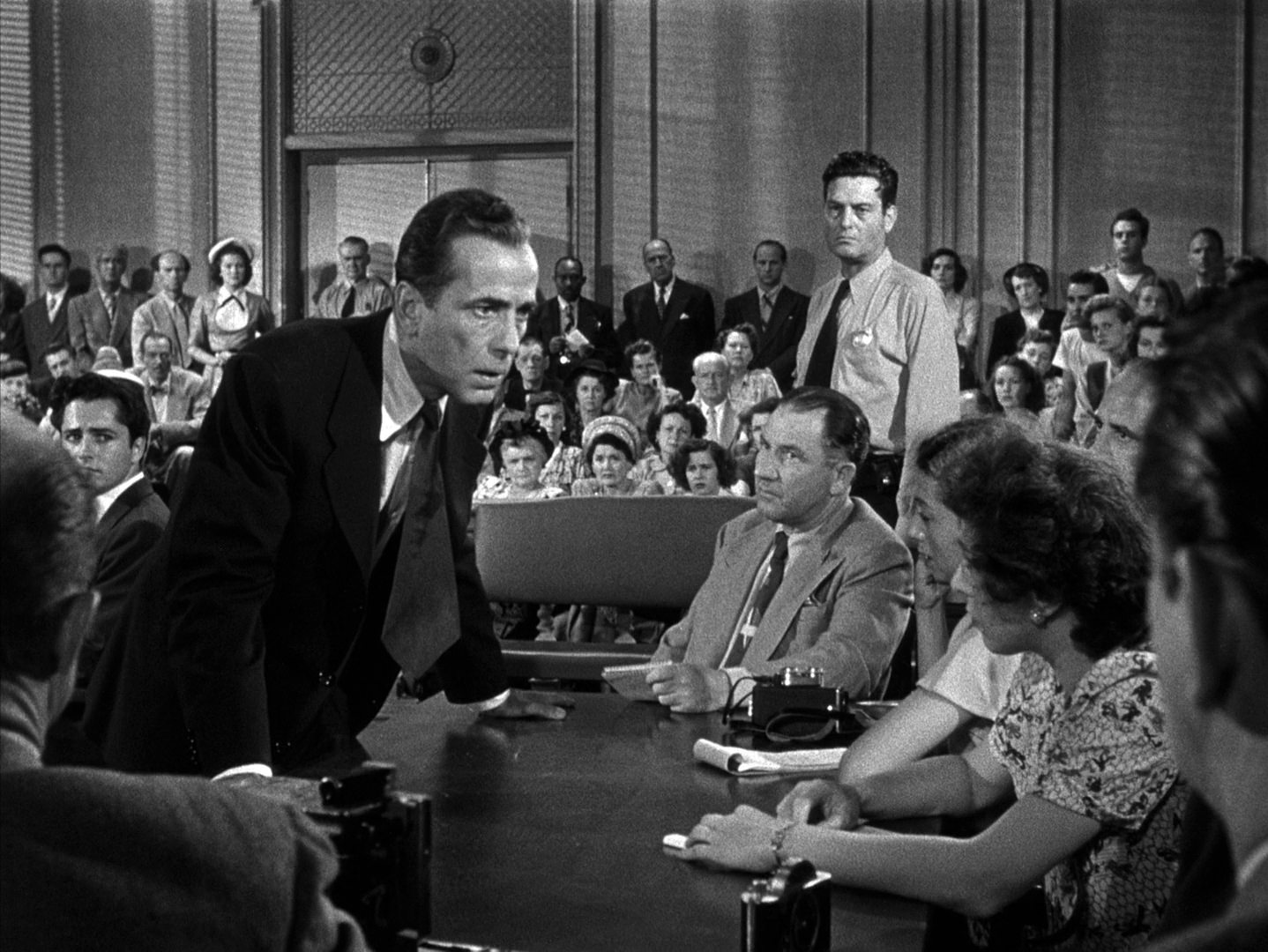 Black and white scene with Humphrey Bogart as a lawyer leaning over a desk in the courtroom during his persuasion.