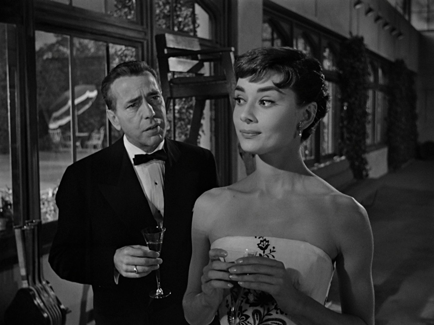 Black and white scene with Humphrey Bogart and Audey Hepburn in a hall; both are wearing evening clothes and are each holding a champagne glass in their hand.