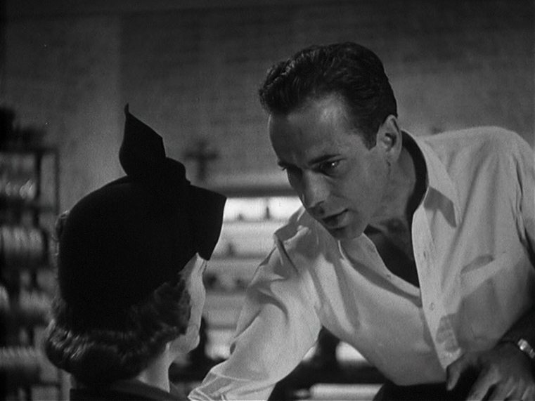 Black and white close-up of Humphrey Bogart in a white shirt: He bends down in conversation to a woman, film reels are stored on a shelf in the background.