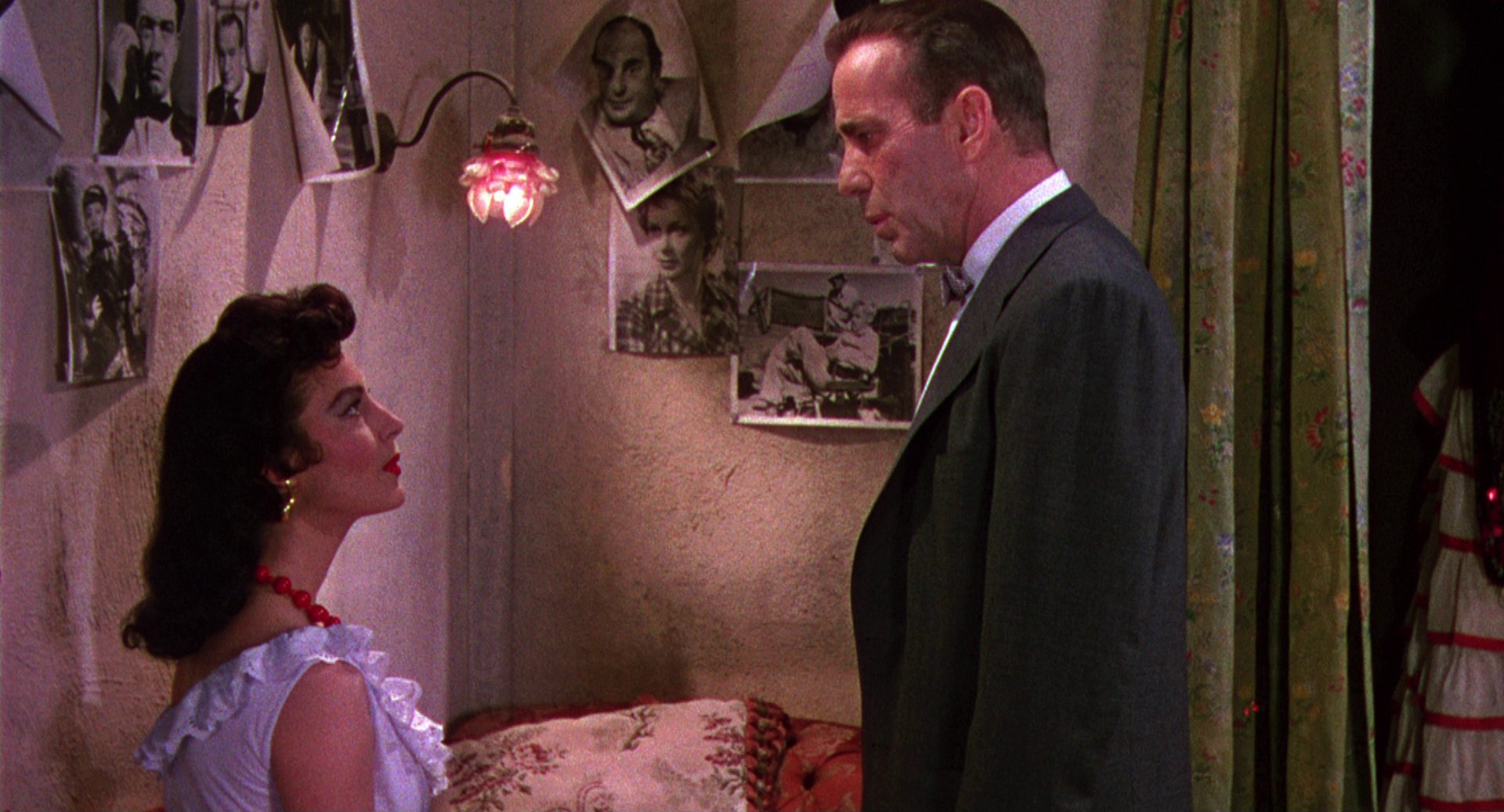 Links im Bild Ava Gardner als Tänzerin in ihrer Garderobe; sie blickt von unten herab zu dem vor ihr stehenden Humphrey Bogart, der einen Hollywoodregisseur im Anzug mit Fliege spielt; an der Zimmerwand hängen mehrere Fotografien von Berühmtheiten.