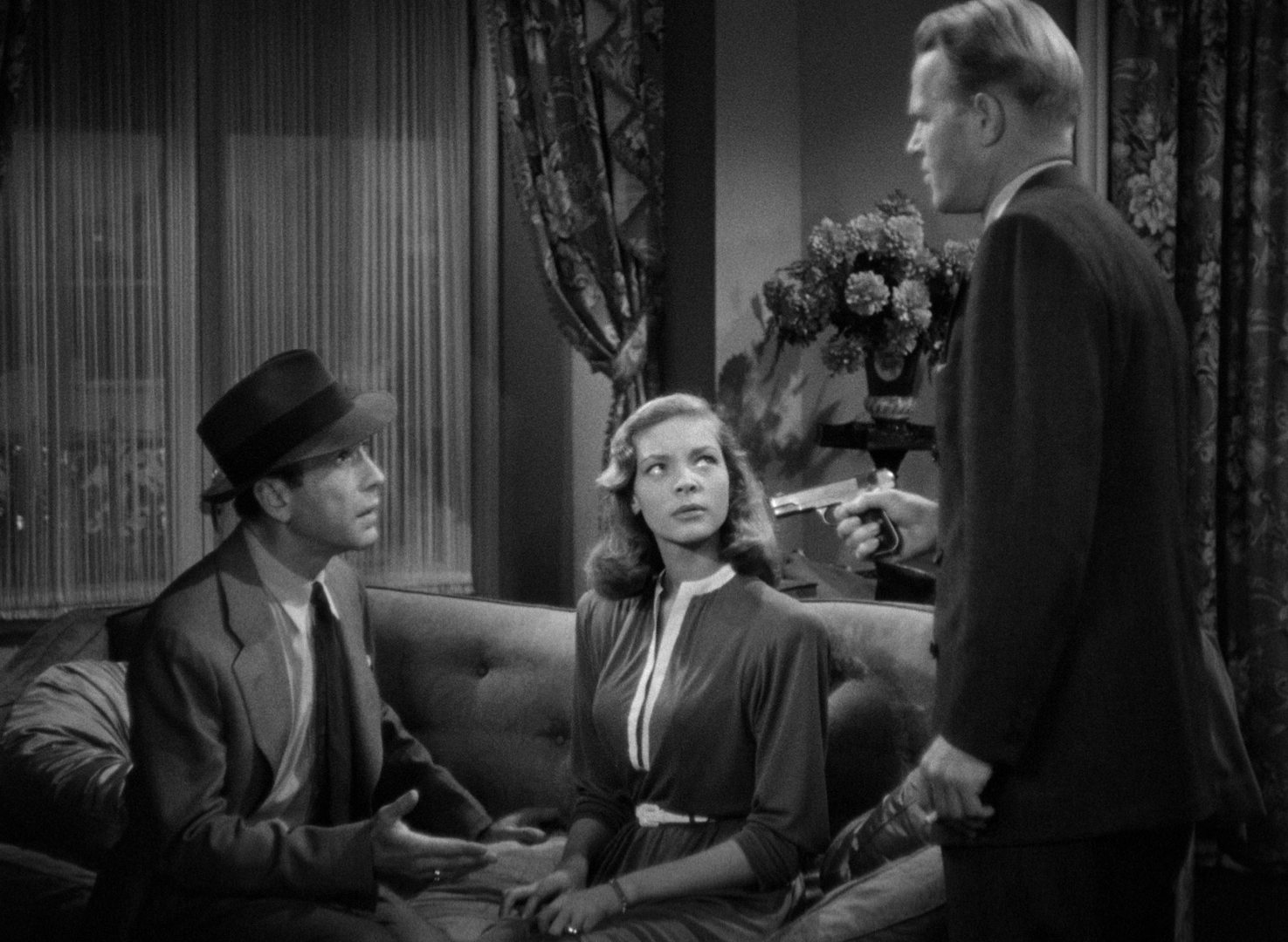 Black-and-white scene with Humphrey Bogart and Lauren Bacall in a flat on a couch, both looking up at a man with a drawn pistol.